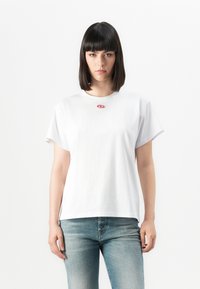 Kvinne med rett, mørkt hår iført en hvit T-skjorte med en liten rosa logo og blå jeans, stående mot en ensfarget hvit bakgrunn.