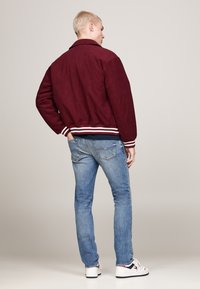 Giacca bomber bordeaux con polsini e orlo a costine, abbinata a jeans di denim blu chiaro e sneakers bianche, vista da dietro.