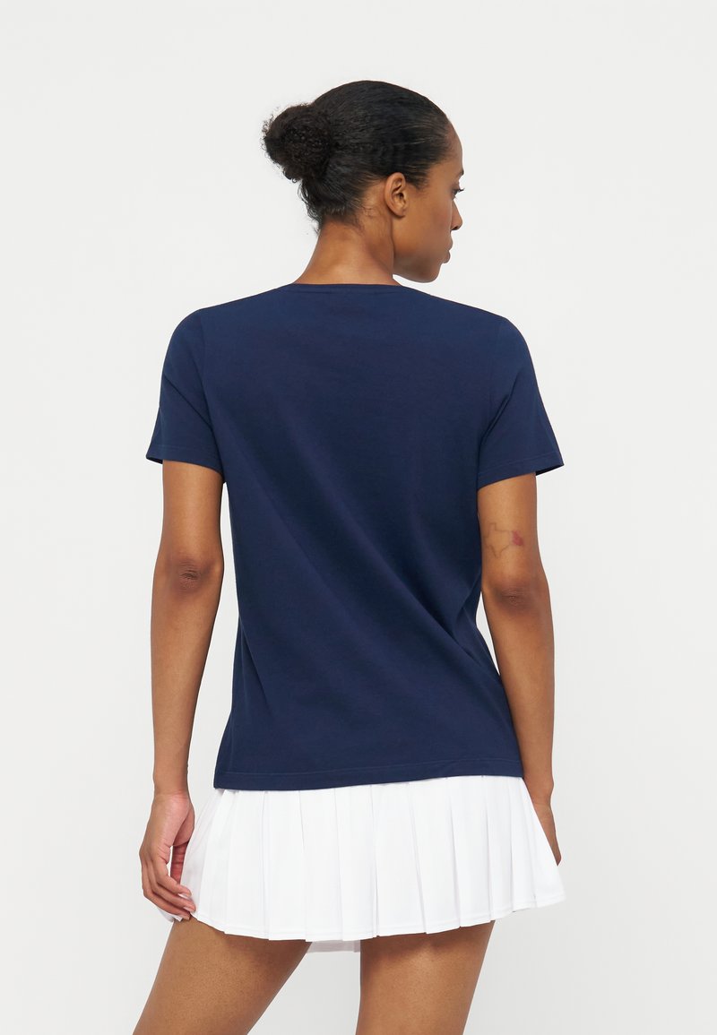 Femme aux cheveux foncés coiffés en chignon, portant une chemise bleu marine à manches courtes et une jupe blanche plissée, tournée dos à la caméra.