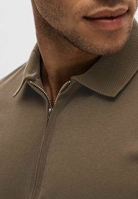 Brun polo skjorta med en texturerad krage, med en silverfärgad dragkedja. Tyget verkar slätt med minimalt synliga sömmar.