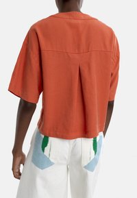 Rdzawoo-pomarańczowa bluzka z krótkim rękawem, wykonana z bawełny, z plisą z tyłu i luźnym krojem. Brak widocznych metek lub akcentów.