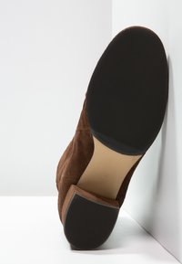 Braune Wildleder-Ankle Boots von hinten gezeigt. Sie verfügen über eine strukturierte schwarze Gummisohle und eine glatte beigefarbene Innensohle.