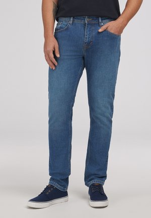 Mężczyzna w niebieskich dżinsach i granatowych trampkach stoi z jedną ręką w kieszeni na jednolitym tle.