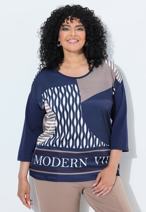 Donna sorridente con capelli ricci neri indossa una maglietta a motivi blu navy, beige e bianco con testo "MODERN VIB" e pantaloni beige, in piedi contro una parete bianca.