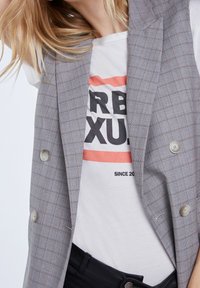 Grå rutig dubbelknäppt väst med ljusa knappar över en vit grafisk t-shirt med fet svart text och ett orange rand. Svarta byxor synliga.