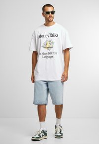 Balta medvilnė t-shirt su grafiniu spaudiniu, vaizduojančiu pinigus ir tekstą "Pinigai kalba daugeliu skirtingų kalbų". Derinama su šviesiais džinsiniais šortais ir žaliais sportbačiais.