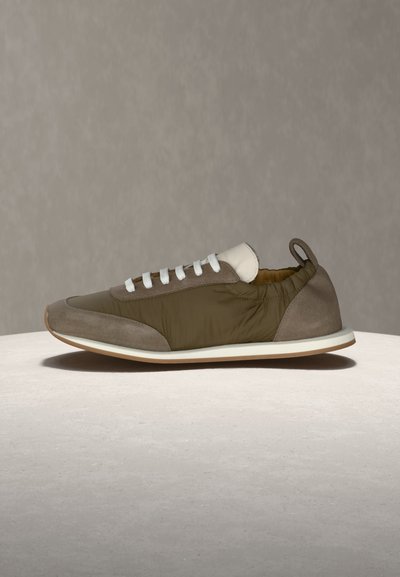 Καφέ και μπεζ αθλητικό παπούτσι με λευκά κορδόνια, ταινία έλξης στη φτέρνα και λευκή σόλα, τοποθετημένο σε φωτεινή επιφάνεια μπροστά σε απλό γκρι φόντο.