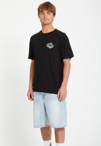 Schwarzes Baumwoll-T-Shirt mit einem weißen Volcom-Logo auf der Brust, kombiniert mit hellblauen Jeansshorts und weißen Socken, vor einem weißen Hintergrund stehend.