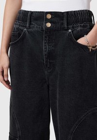 Pantaloni in denim nero con vita elasticizzata e due bottoni dorati, tasche laterali e una vestibilità rilassata con dettagli di cucitura distintivi.