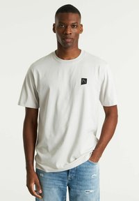 Lichtgrijze t-shirt van katoen, met een ronde hals, korte mouwen en een klein zwart logo op de borst. Draagt bij blauwe versleten jeans.
