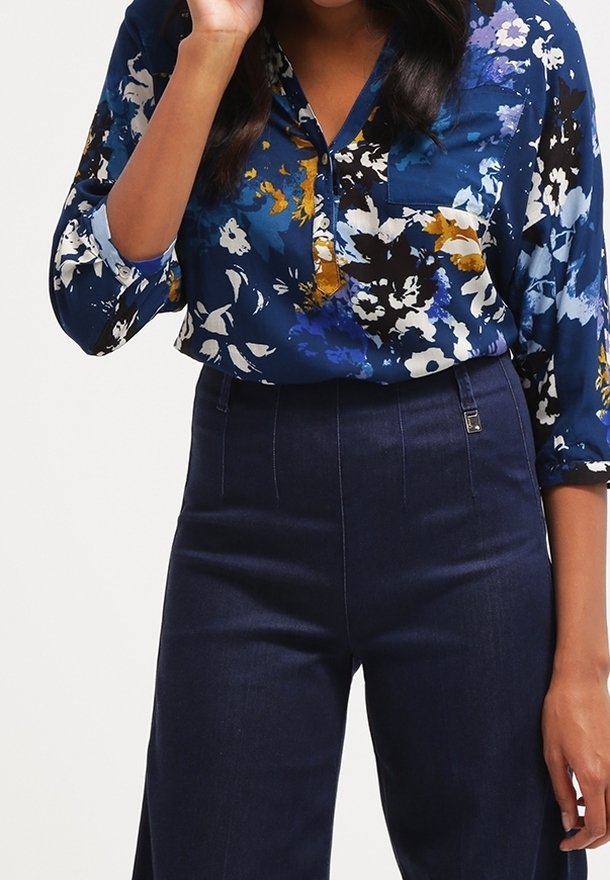 Femme portant un chemisier à fleurs bleu avec une poche poitrine rentrée dans un pantalon bleu foncé taille haute, debout avec une main près du visage.