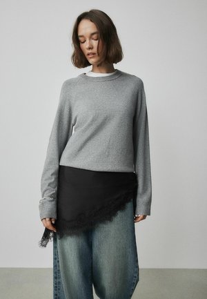 Grå långärmad tröja med bred urringning, ovanpå en svart kjol med spetsdetaljer och lösa jeansbyxor med fransiga kanter.