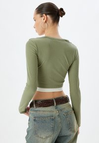 Olivgrünes, geripptes, langärmliges Cropped-Top mit weißem Saum, kombiniert mit hoch sitzenden hellblauen Jeans und einem geflochtenen braunen Ledergürtel.