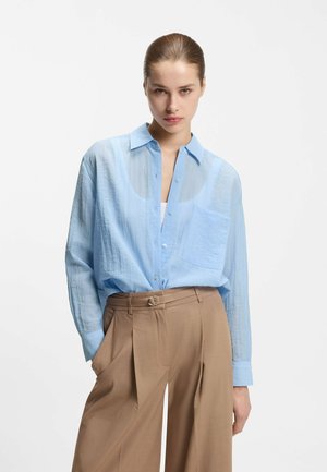 Kobieta nosząca jasnoniebieską przezroczystą koszulę zapinaną na guziki na białym topie oraz szerokie, wysokie spodnie w kolorze beżowym z paskiem z pierścieniem D.