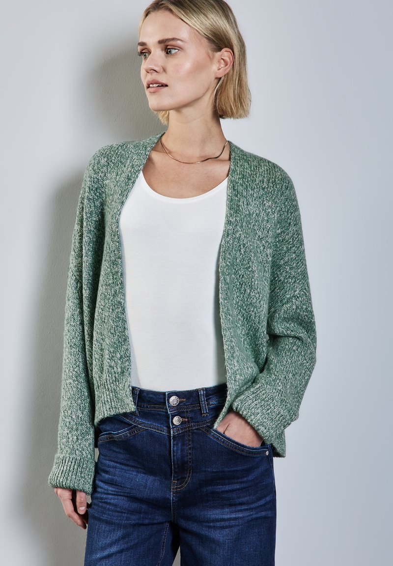 Cardigan en maille vert clair avec un devant ouvert, manches texturées, associé à un top blanc et un jean bleu foncé avec des boutons doubles.