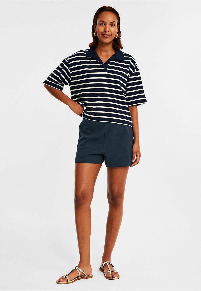 Laivaston sininen ja valkoinen raidallinen lyhythihainen polo-paita, joka on yhdistetty tummansinisiin shortsikaloihin ja litteisiin sandaaleihin. Asu on rentoa leikkuuta ja ribbitekstuuri. 