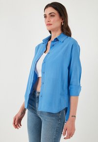 Camicia blu chiaro con bottoni realizzata in un tessuto liscio; presenta un colletto, maniche lunghe arrotolate e un orlo arrotondato.