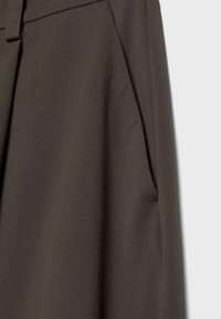 Gros plan sur un pantalon en tissu marron foncé montrant la poche de couture latérale et une partie de la ceinture sur un fond clair.