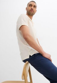 Polo shirt beige chiaro a maniche corte, con colletto classico e spacchi laterali, abbinato a pantaloni slim-fit blu scuro.