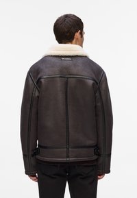 Chaqueta de borrego marrón con un cuello blanco, ribetes de cuero negro y correas ajustables en la cintura. Diseño liso en la parte trasera, con mínima marca.
