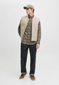 Beige ærmeløs vest over en brun ternet skjorte, mørke jeans og lysebrune støvler. Kappen matcher vestens farve, hvilket skaber et koordineret look.