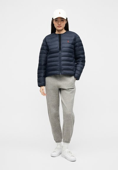 Γυναίκα που φοράει λευκό καπέλο, σκούρο μπλε μπουφάν puffer, γκρι φόρμα, λευκές κάλτσες και λευκά αθλητικά παπούτσια, στέκεται μπροστά σε απλό φόντο.