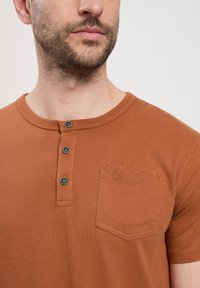 Brązowa koszulka henley z krótkim rękawem, z kieszonką, wyposażona w trzy ciemne guziki i gładką strukturę. Kołnierz jest zaokrąglony i ma przeszywaną krawędź.