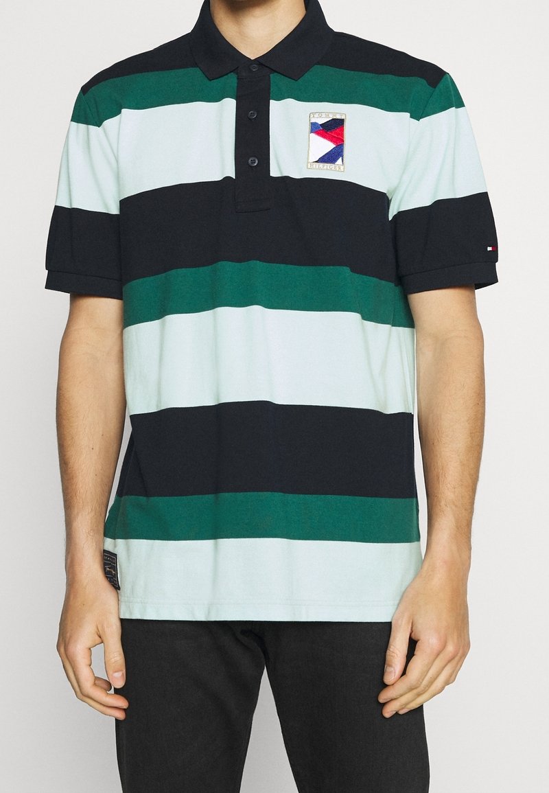 Homme portant un polo à manches courtes avec des rayures horizontales noires, vertes et bleu clair, et un patch de logo sur la poitrine gauche.