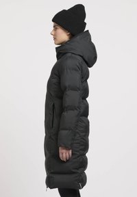 Cappotto lungo imbottito nero con cappuccio, caratterizzato da un esterno liscio, tasche laterali e un interno foderato, abbinato a un berretto a coste nero.