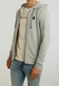 Hombre vistiendo una sudadera con cremallera gris claro sobre una camiseta beige con el texto "CHASE", combinado con jeans azul claro, de pie frente a un fondo sencillo.
