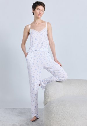 Vrouw staat op blote voeten met één been op een wit gestructureerde bank, gekleed in een witte bloemige pyjama met kanten afwerking aan het hemdje.