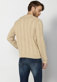 Maglione beige a maglia con collo a giro e polsini a costine, abbinato a jeans in denim blu scuro. Trama a maglia con motivi in rilievo.