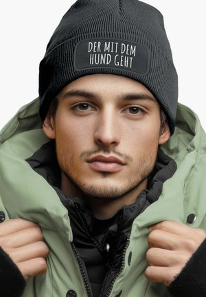 Black knitted beanie with a rectangular patch reading "DER MIT DEM HUND GEHT," worn with a light green padded jacket with a black inner lining.
