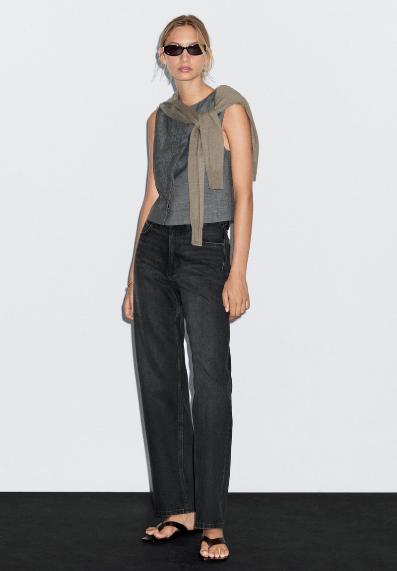 Graues ärmelloses Top mit einem beigen Pullover, der über die Schultern drapiert ist, kombiniert mit hochgeschnittenen schwarzen geraden Jeans und schwarzen Sandalen.
