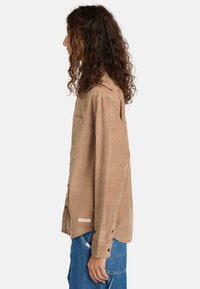 Beige Cordhemd mit Knopfleiste, seitlicher Tasche, lässigem Schnitt und langen Ärmeln, kombiniert mit blauen Jeans.