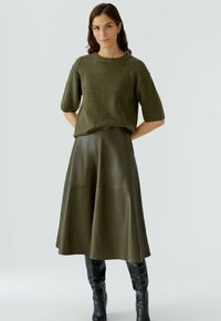 Olivgrüner, gestrickter Pullover mit kurzen Ärmeln, kombiniert mit einem passenden, knielangen Ledrock, ergänzt durch figurbetonte, schwarze Stiefel bis zum Knie.
