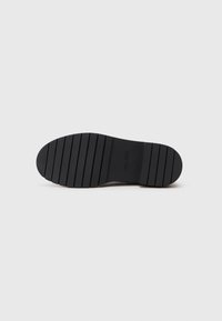 Suela de zapato de goma negra con una superficie texturizada, que presenta surcos horizontales para un mejor agarre y una forma de punta redondeada. Marcada con "ALDO."