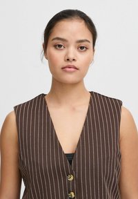 Gilet sans manches marron avec des rayures verticales blanches, décolleté en V plongeant et deux boutons dorés à l'avant. Tissu texturé avec une coupe ajustée.