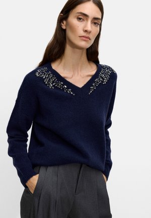 Granatowy sweter dzianinowy z dekoltem w kształcie V i ozdobnymi ramionami, na których znajdują się florystyczne wzory wykonane z metalicznej nici i kamieni szlachetnych.
