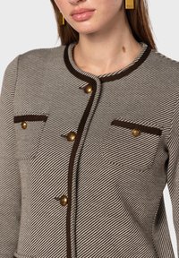 Texturerad beige och brun kavaj med guldfärgad knappar, rund halsringning, två bröstfickor och kontrasterande bruna detaljer.