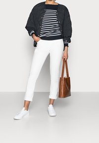 Veste bomber noire matelassée portée sur un pull rayé bleu marine et blanc, associée à un pantalon blanc retroussé et des baskets blanches, tenant un sac fourre-tout marron.
