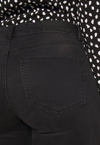 Vaqueros de mezclilla negra con un leve estiramiento, con una cintura de tiro medio, bolsillos traseros y una textura suave. Etiqueta visible cerca de la cintura.