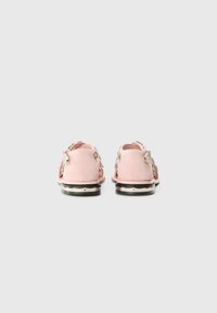 Pink suede slip-on shoes with a low heel, featuring metallic accents and decorative hardware on the sides. Smooth texture and rounded shape.