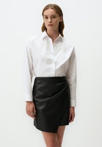 Chemise blanche à manches longues, accompagnée d'une jupe en simili cuir noire à enrouler, présentant une texture lisse et un design diagonal.