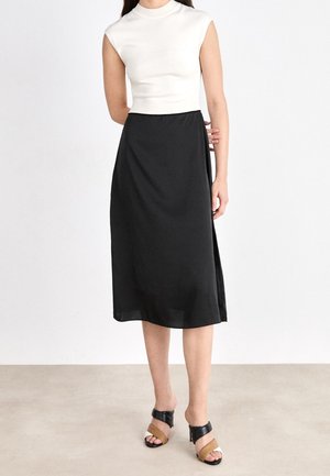 Kvinde iført en hvid ærmeløs top, sort midi-nederdel og tofarvede åbne hælede sandaler, stående mod en ensfarvet hvid baggrund.