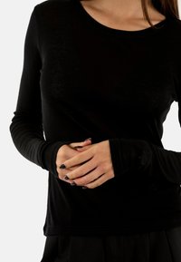 Donna che indossa una maglietta nera a maniche lunghe con fori per i pollici, mani incrociate in vita, in piedi contro uno sfondo bianco.