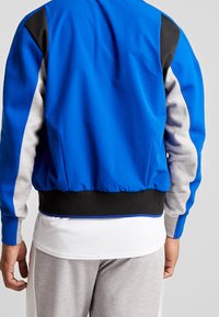 Chaqueta bomber azul con mangas grises y acentos negros. Cintura negra acanalada, textura suave, diseñada con secciones en bloque de color.