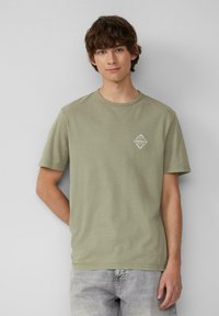 Oliwkowa bawełniana koszulka z krótkim rękawem, okrągłym dekoltem i małym białym logo na piersi. Gładka struktura, luźny krój.