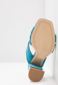 Korkeakorkoiset teal-väriset mokkatossut, joissa on avotossu ja hopeiset niitit. Litteä, beige kumipohja, jossa on kolmionmuotoisia kuvioita.