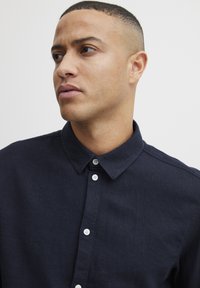Camisa oscura de lino con botones, de color azul marino, que presenta un cuello, placket con botones blancos y una tela texturizada. Vista en primer plano.